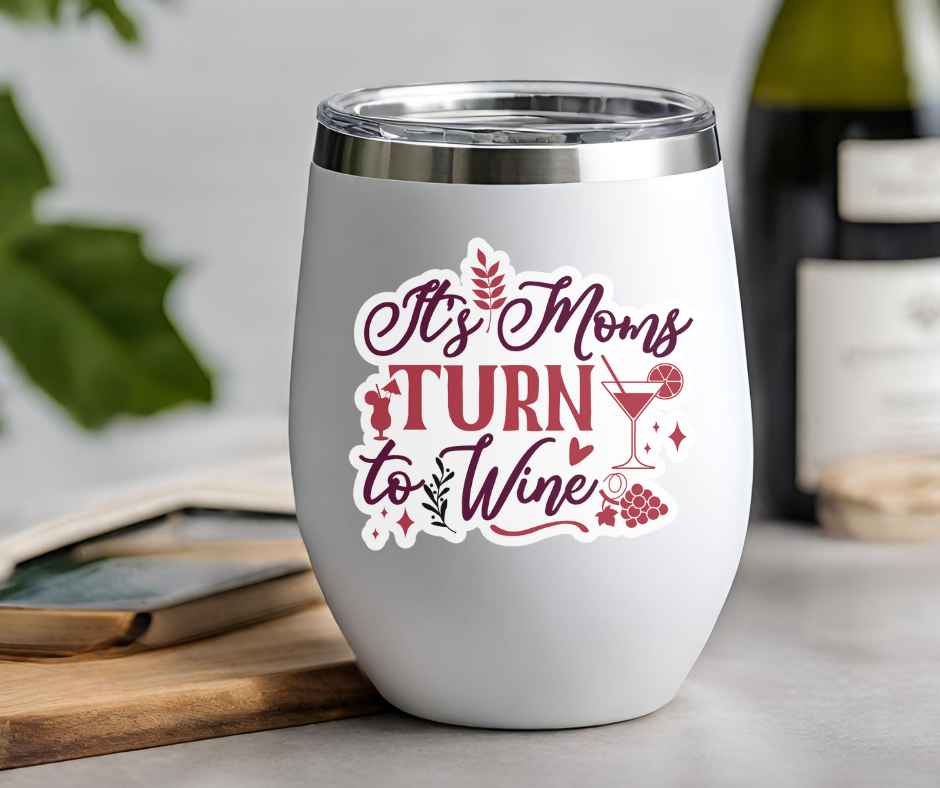 Insulated wine tumbler with "It's Moms Turn to Wine" text design on a countertop with wine bottle and greenery background.