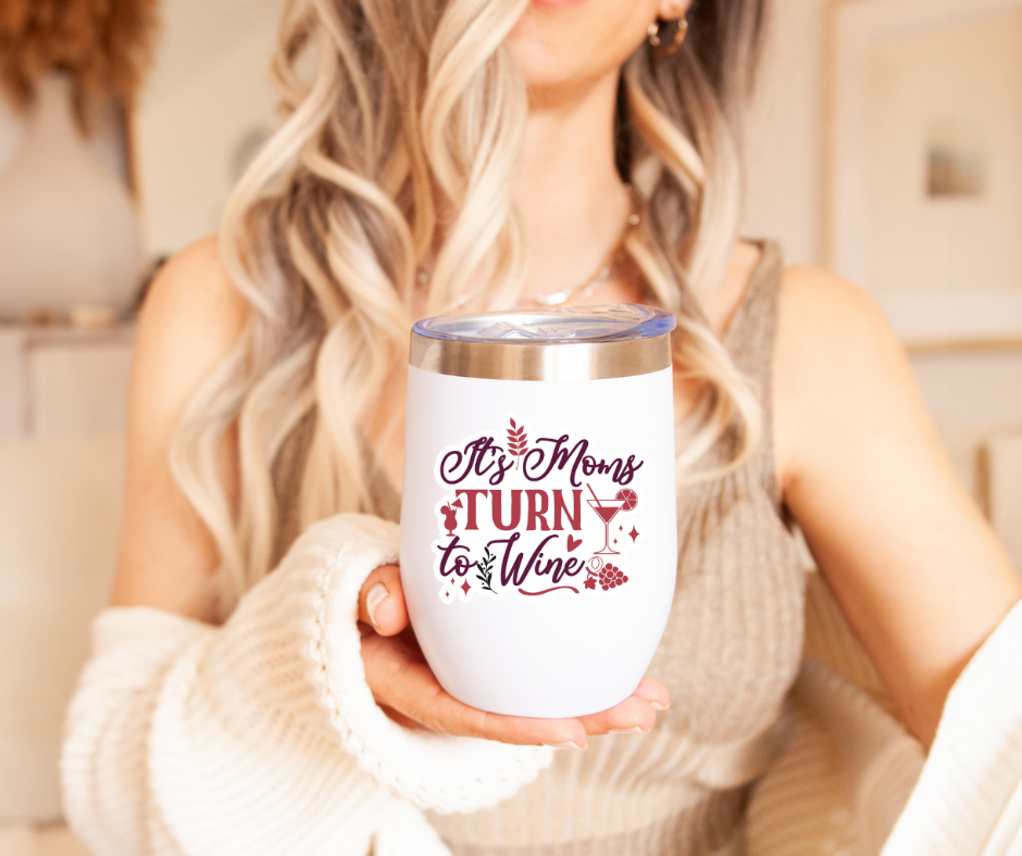 Woman holding a white insulated wine tumbler with the text "It's Mom's Turn to Wine" in a cozy indoor setting.