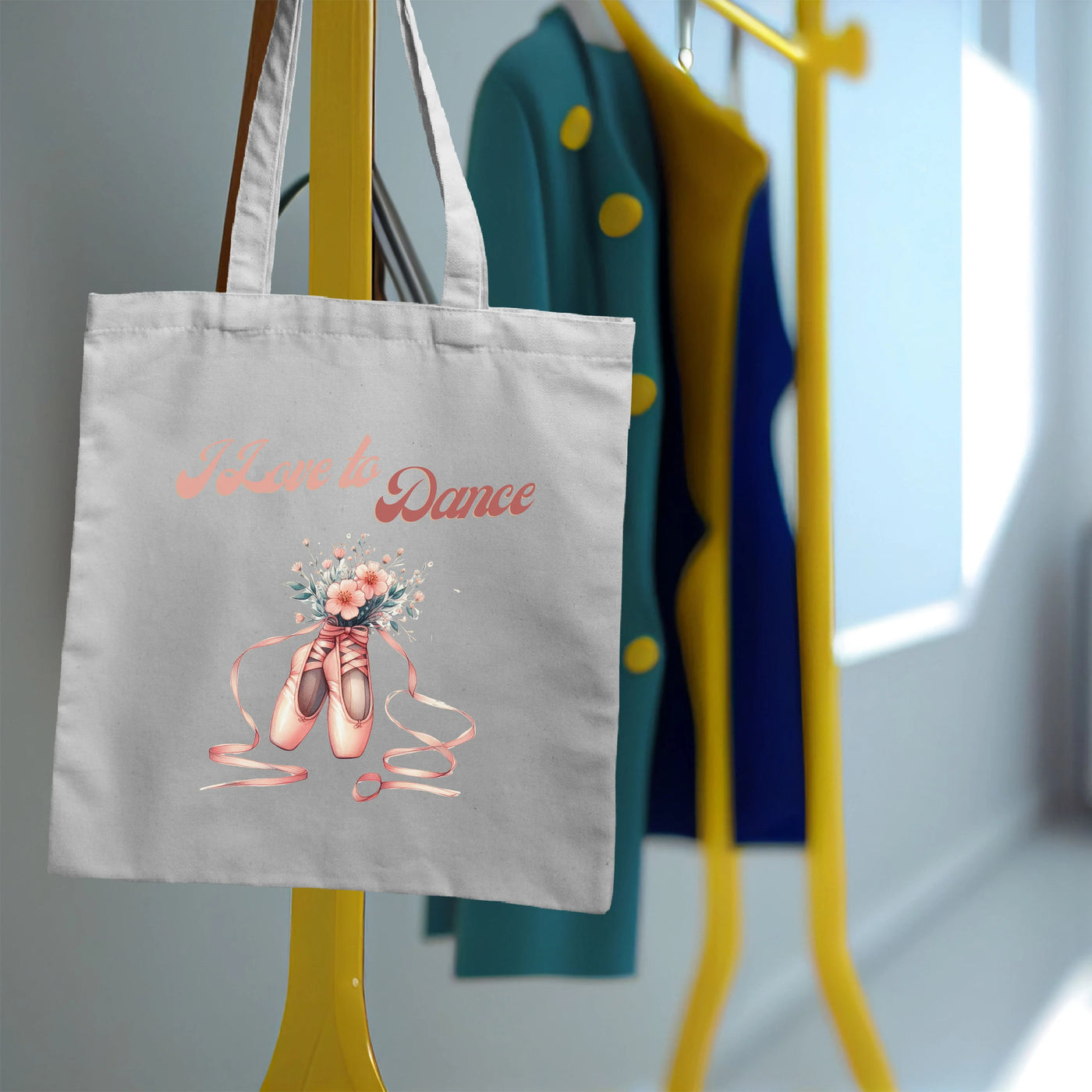 Tote bag with ballet shoes and flowers design hanging on a yellow rack, with colorful coats in the background.