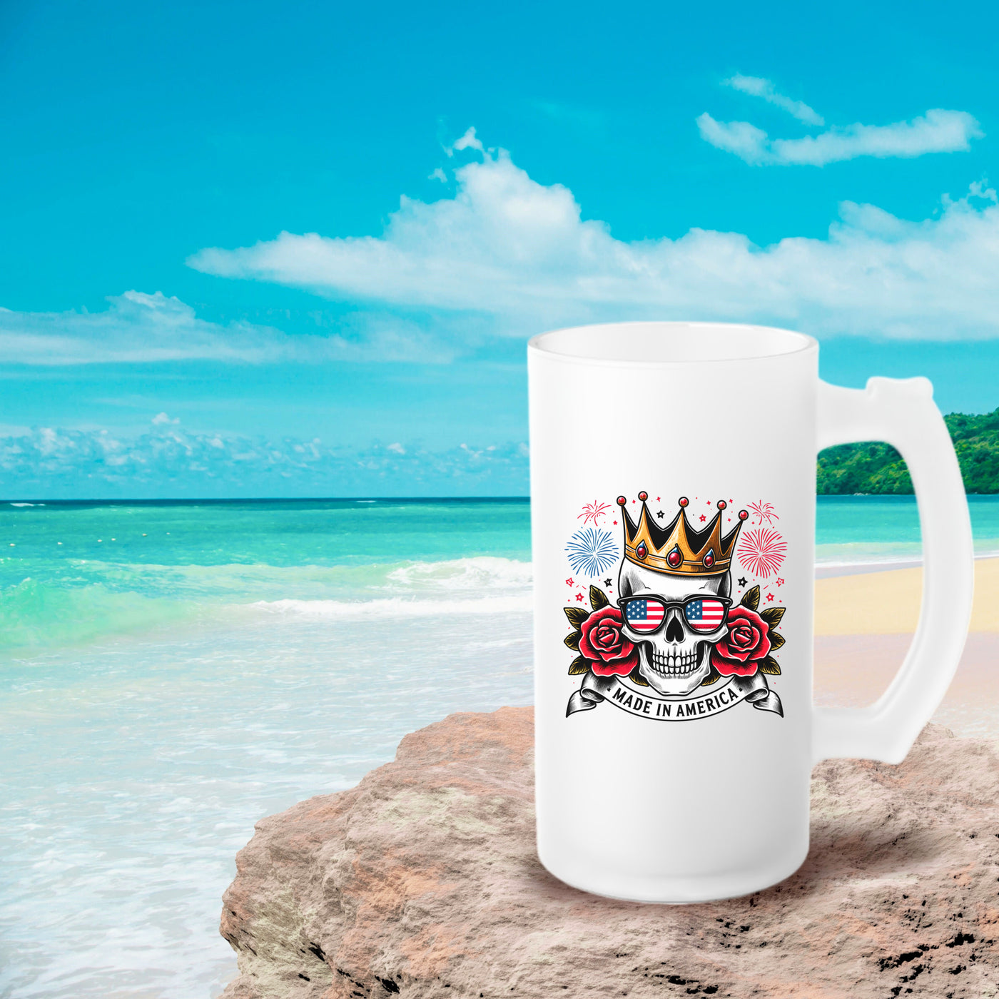 White mug with a skull, roses, and crown design saying "Made in America" placed on a beach rock with ocean and sky background.