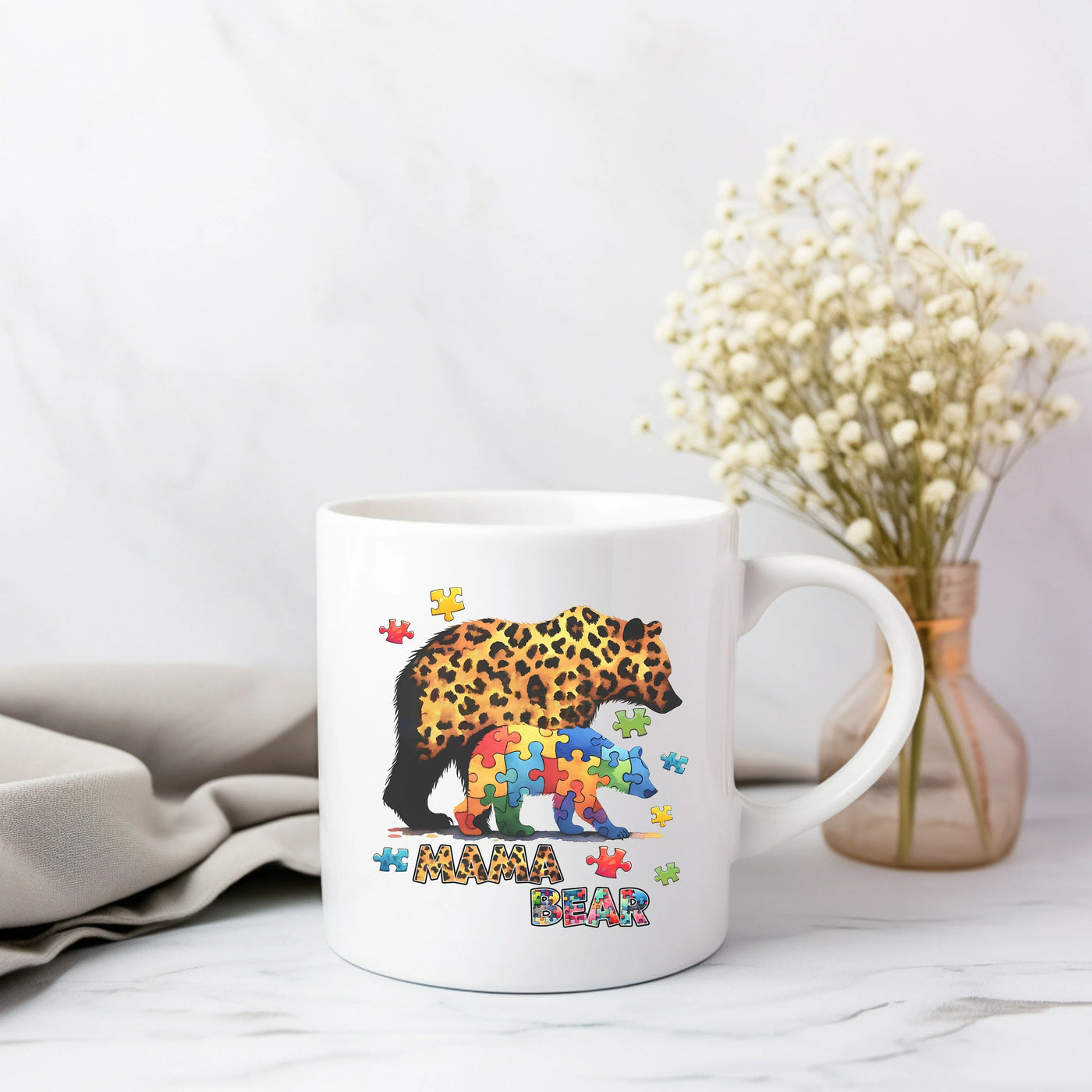 White coffee mug with "Mama Bear" and puzzle design, next to a vase with flowers, on a marble surface.