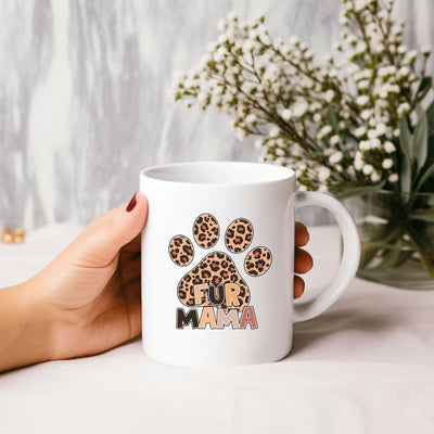 Hand holding white mug with leopard print paw and "Fur Mama" text, with flowers in the background