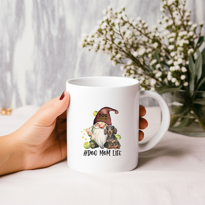 Hand holding a white mug with "Dog Mom Life" design featuring a gnome and dog.