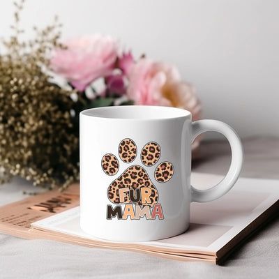 White coffee mug with leopard print paw print design and text "Fur Mama," on a stack of books with flowers in the background.