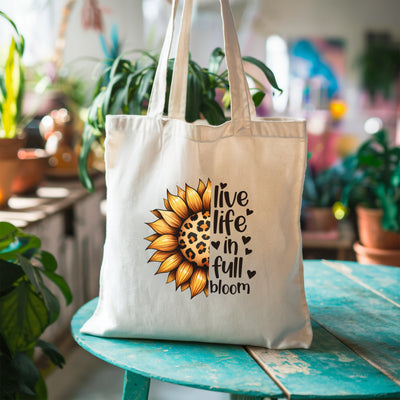 White canvas tote bag with sunflower and leopard print design, text reading "Live life in full bloom," displayed on a green wooden table.