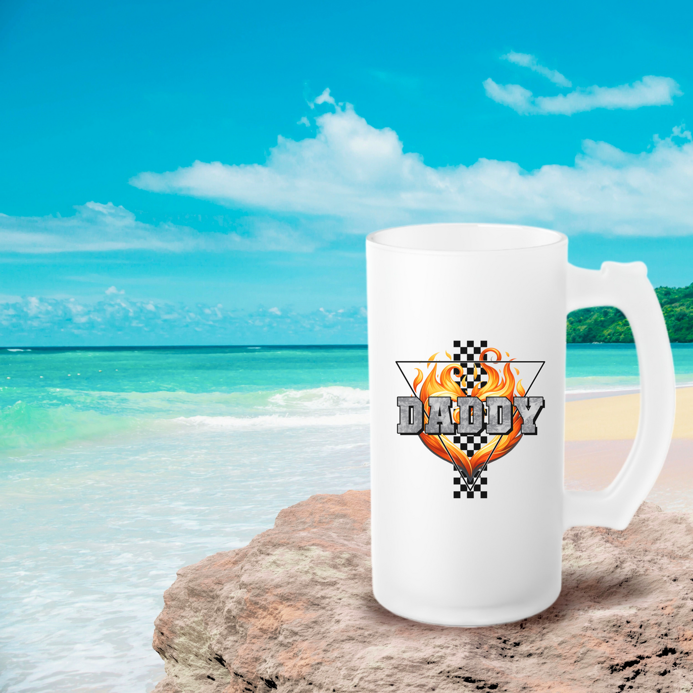 White mug with a fiery design and the text "DADDY", placed on a rock by the beach with a calm blue ocean and cloudy sky in the background