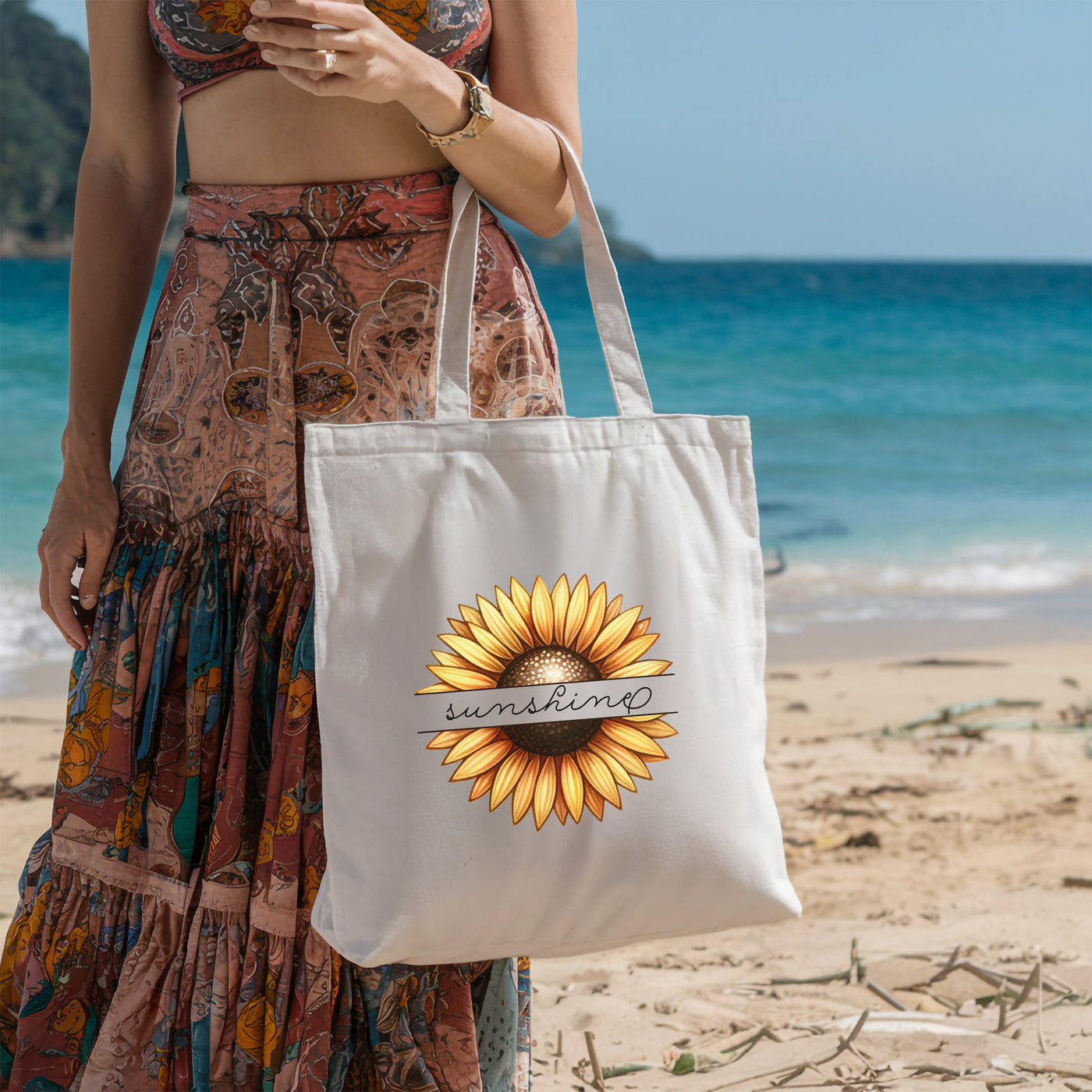 Woman holding white tote bag with sunflower design and "sunshine" text on a beach.