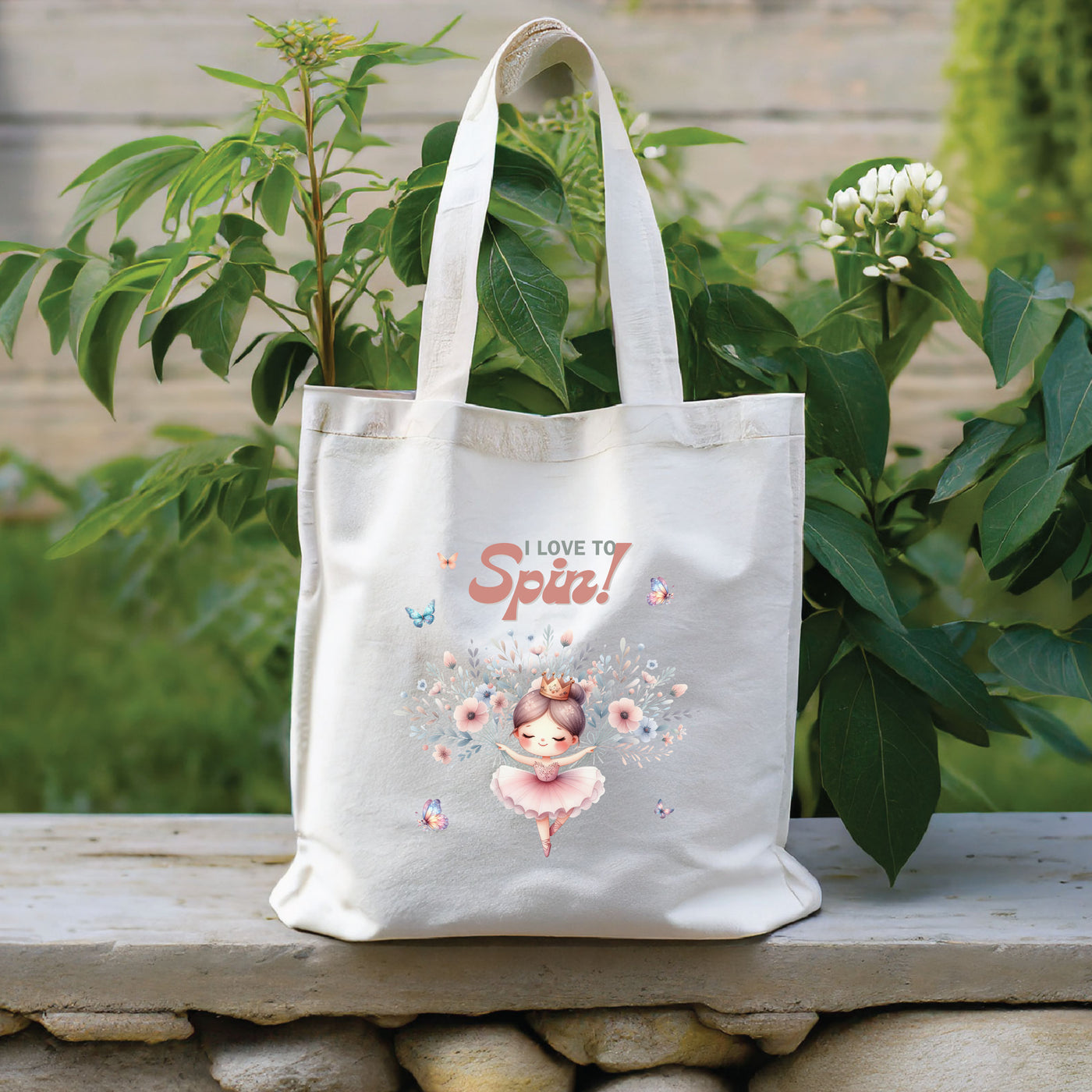 White tote bag with "I Love to Spin!" design featuring a dancing girl, flowers, and butterflies, placed on an outdoor stone surface.