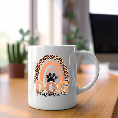 White "Dog Mama" coffee mug with paw print design and leopard print rainbow on wooden desk near plants and laptop.