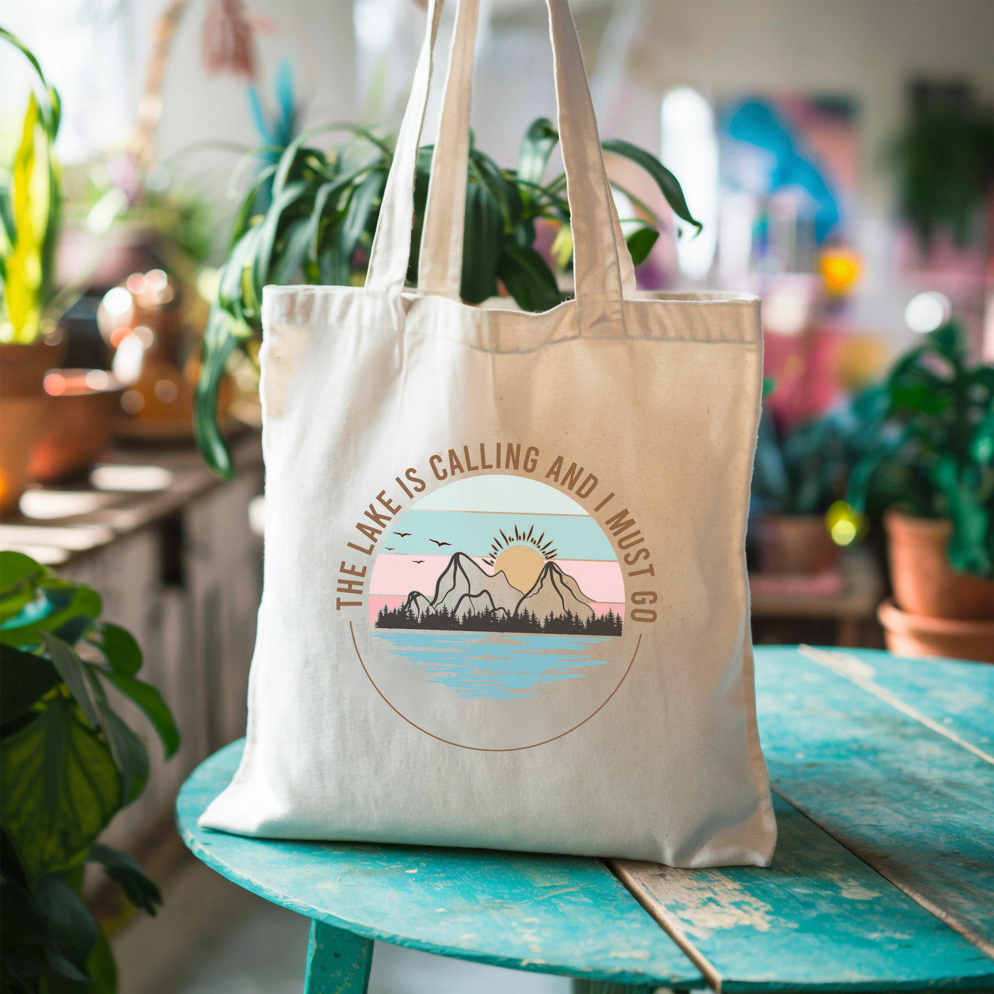 Tote bag with "The Lake is Calling and I Must Go" design featuring mountains and lake, placed on a green wooden table amidst plants