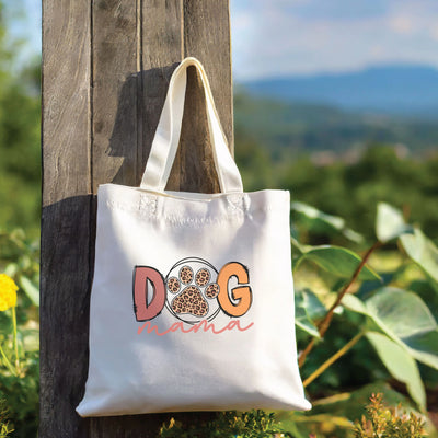 White tote bag with "Dog Mama" text, featuring a paw print design, hanging on a wooden post with a scenic landscape background.