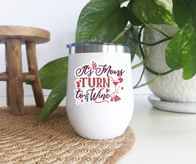 White wine tumbler with "It's Mom's Turn to Wine" text next to greenery and wooden stool for Mother's Day.