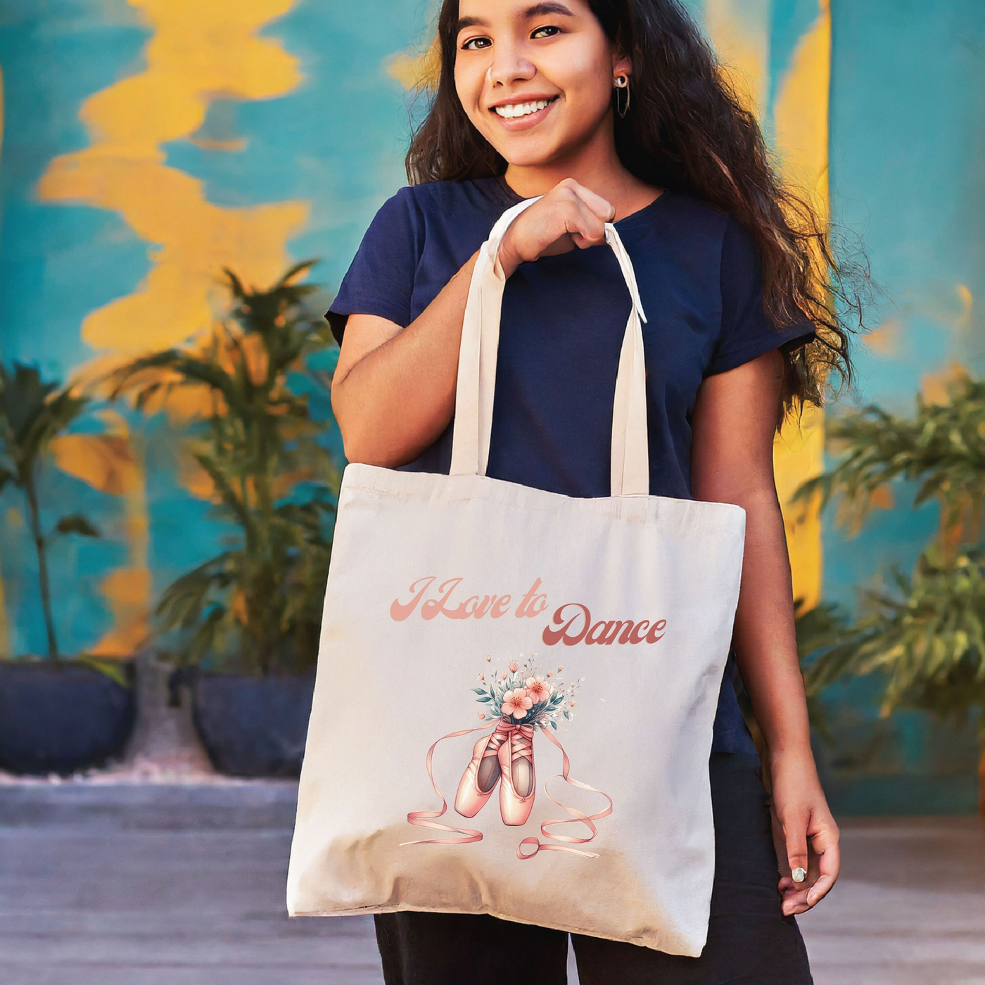 Woman smiling with a beige tote bag that says "I Love to Dance" in cursive text against a colorful wall background.