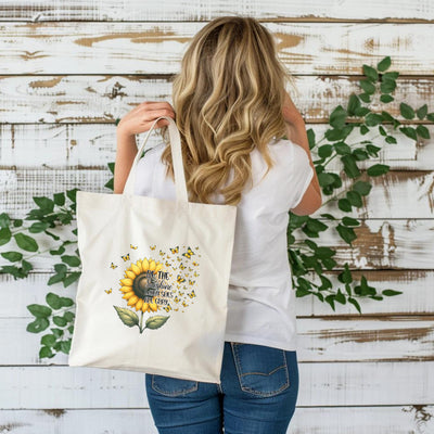 Woman holding a white tote bag with sunflower design and motivational quote against rustic wood and greenery background.