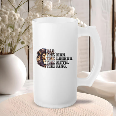 White ceramic mug with lion graphic and text "Dad. The Man. The Legend. The Myth. The King." on a wooden table with decorative flowers in the background.