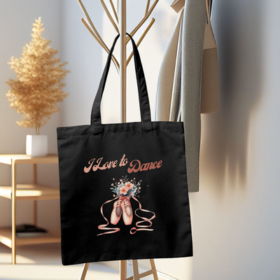 Black tote bag with "I Love to Dance" text and ballet shoes design hanging on a wooden coat rack in a modern, minimalistic room