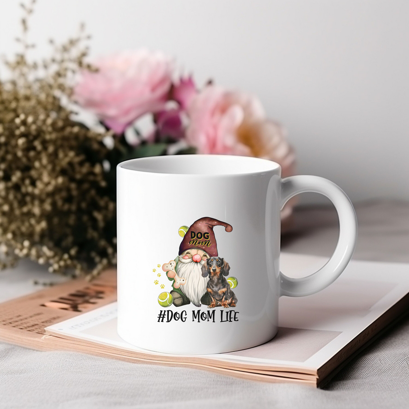 White coffee mug with "Dog Mom Life" text and gnome graphic, placed on books with blurred flowers in the background