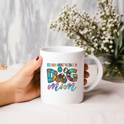 Hand holding a white mug with colorful text "Kinda Busy Being a Dog Mom" next to a floral arrangement