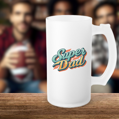 White frosted beer mug with "Super Dad" text, placed on a table with blurred background of people holding a football