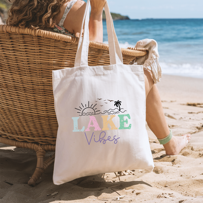 a woman sitting in a chair on the beach with a lake vibe tote bag