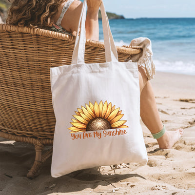 a woman sitting in a chair on the beach with a sunflower tote bag